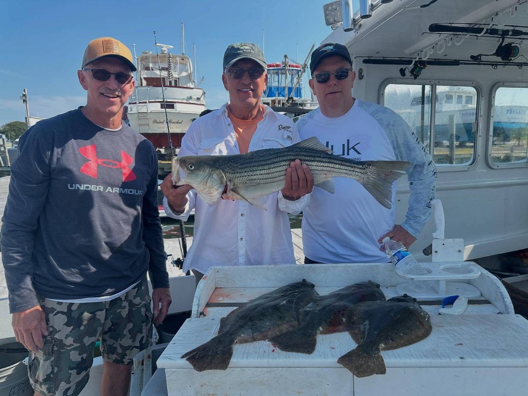 Three adult anglers with striped bass and fluke beside the Anna Maria cabin