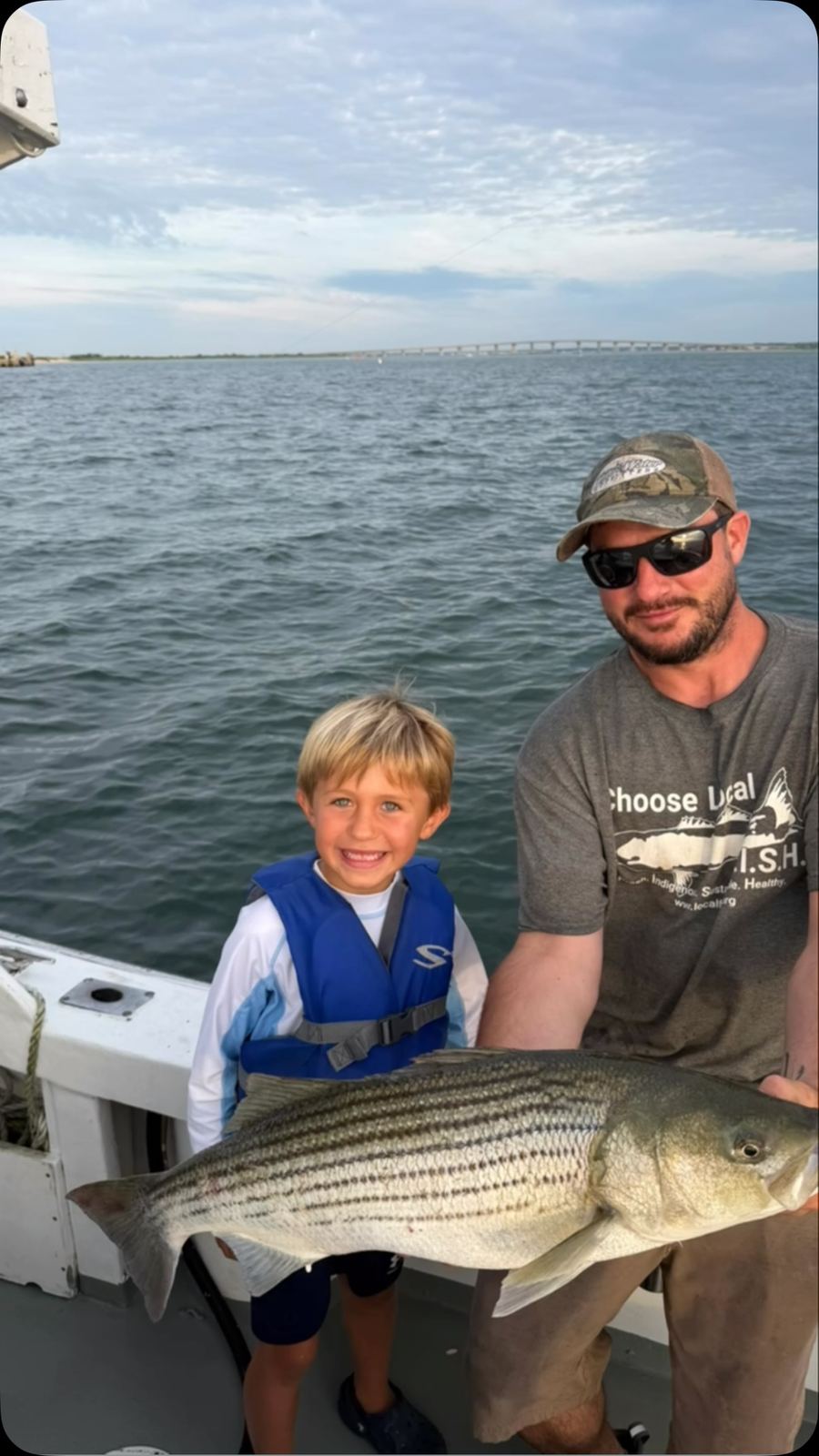 A young child in a life vest standing beside an adult holding a large striped bass