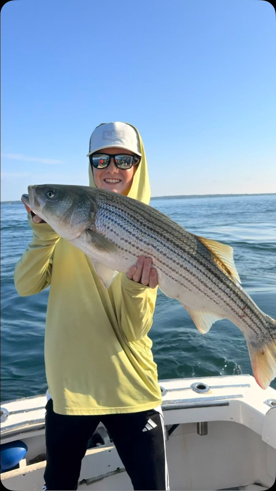 An angler in a yellow hoodie holding a striped bass over calm water